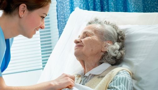 Pflegekraft hält die Hand einer älteren Frau, die im Bett liegt, im hellen Zimmer mit Fensterläden.