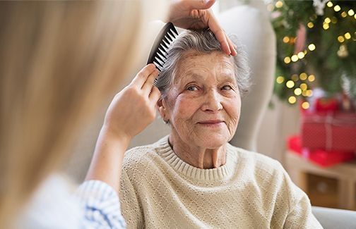 Ältere Person mit grauen Haaren trägt hellen Pullover und wird von einer anderen Person mit Kamm im Haar frisiert, im Hintergrund Weihnachtsbeleuchtung und Geschenke.