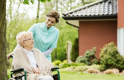 Pflegekraft in türkisfarbener Kleidung schiebt ältere Person mit hellen Haaren und beigem Strickponcho im Rollstuhl im Garten eines Hauses.