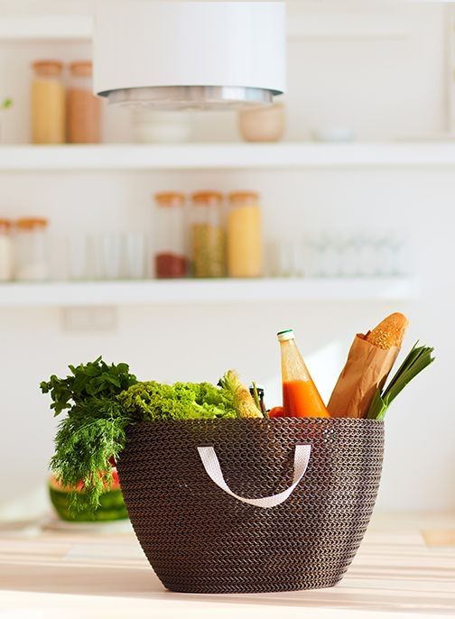 Braune Einkaufstasche mit gewebter Struktur auf Küchentheke, gefüllt mit grünem Salat, Karotten, Baguette und Flasche mit oranger Flüssigkeit.